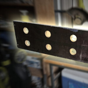 The long wooden board being held in a vice. It has 8 holes drilled in an even 2x4 grid. It's stylised to dramatically highlight the board against the background.