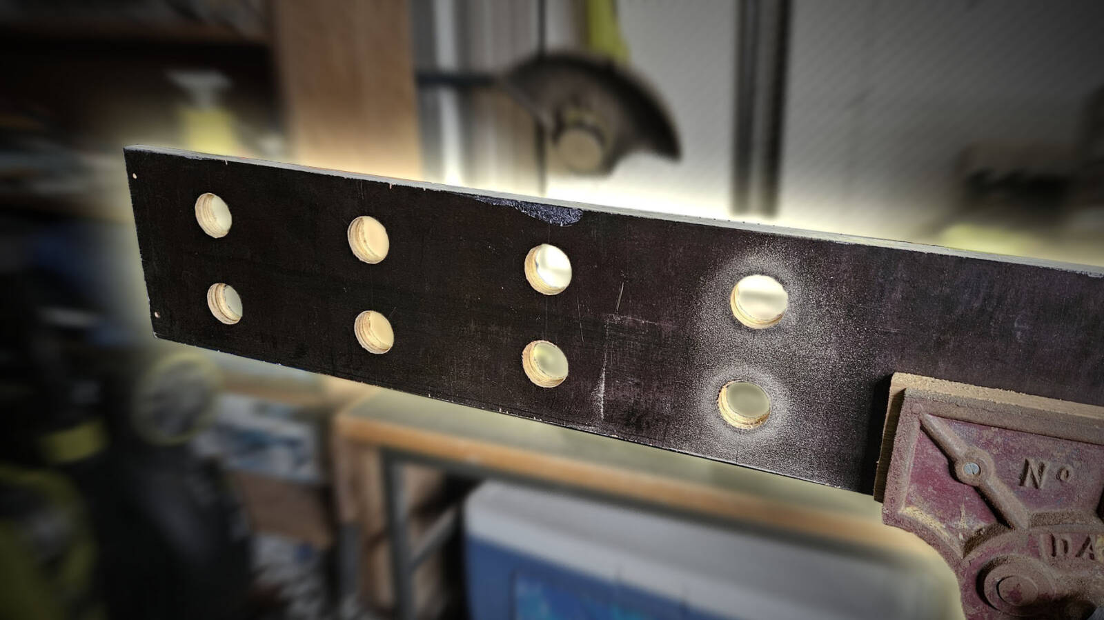 The long wooden board being held in a vice. It has 8 holes drilled in an even 2x4 grid. It's stylised to dramatically highlight the board against the background.